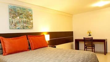 A hotel room with a bed, two orange pillows, a nightstand, a desk with a chair, and a painting on the wall.