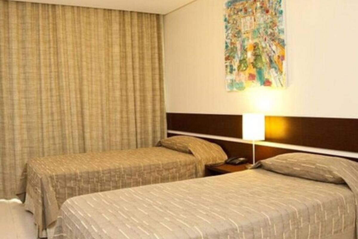 Camera d'albergo con due letti singoli, una testiera, un comodino con una lampada e una tenda.