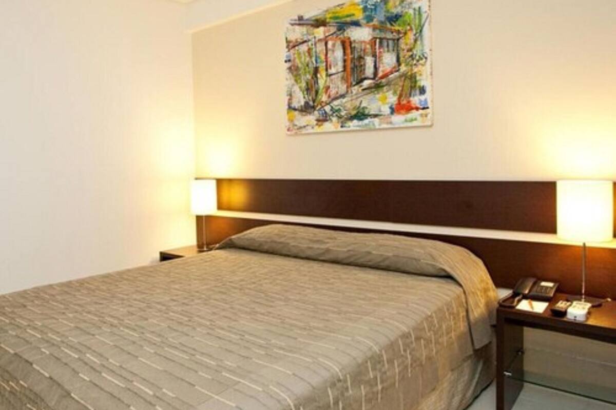 Una camera d'albergo con un letto grande, una testiera, due comodini, una lampada e un quadro appeso al muro.