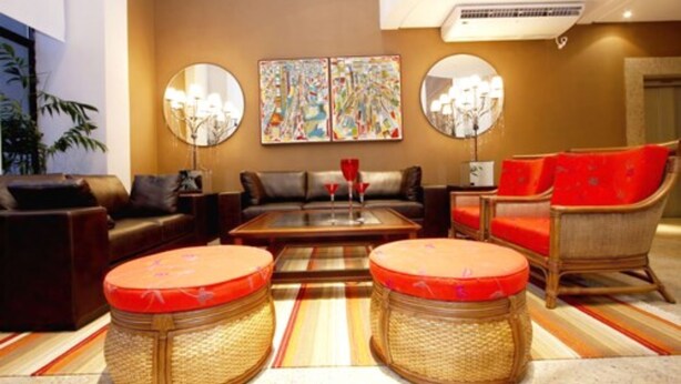 A modern living room with a sofa, armchairs, a coffee table, and two wicker ottomans. There are two round mirrors on the wall and a colorful abstract painting above the coffee table. - América Towers Hotel (Salvador)