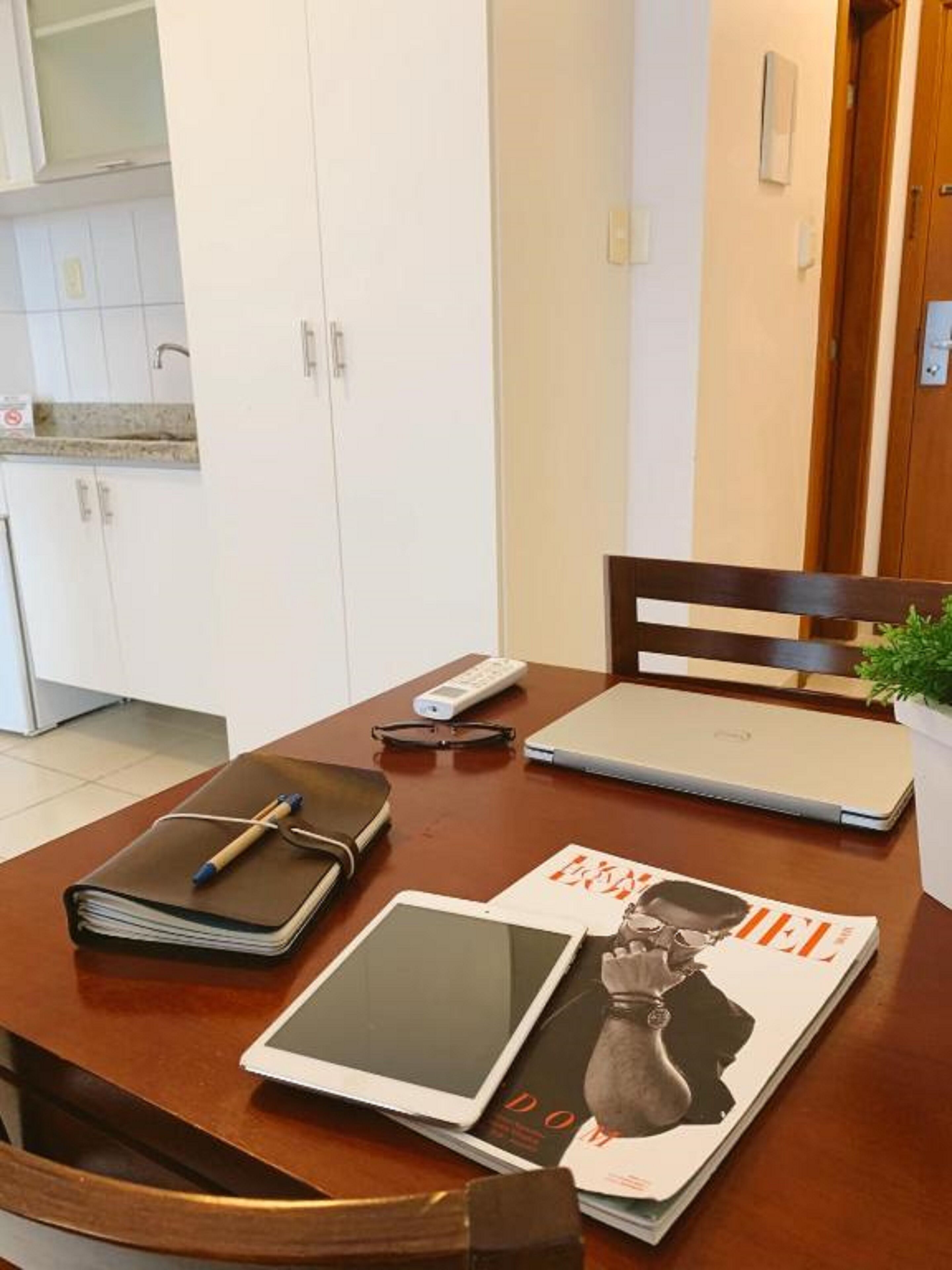 A wooden dining table with a laptop, a tablet, a remote control, a notebook, and a pen.