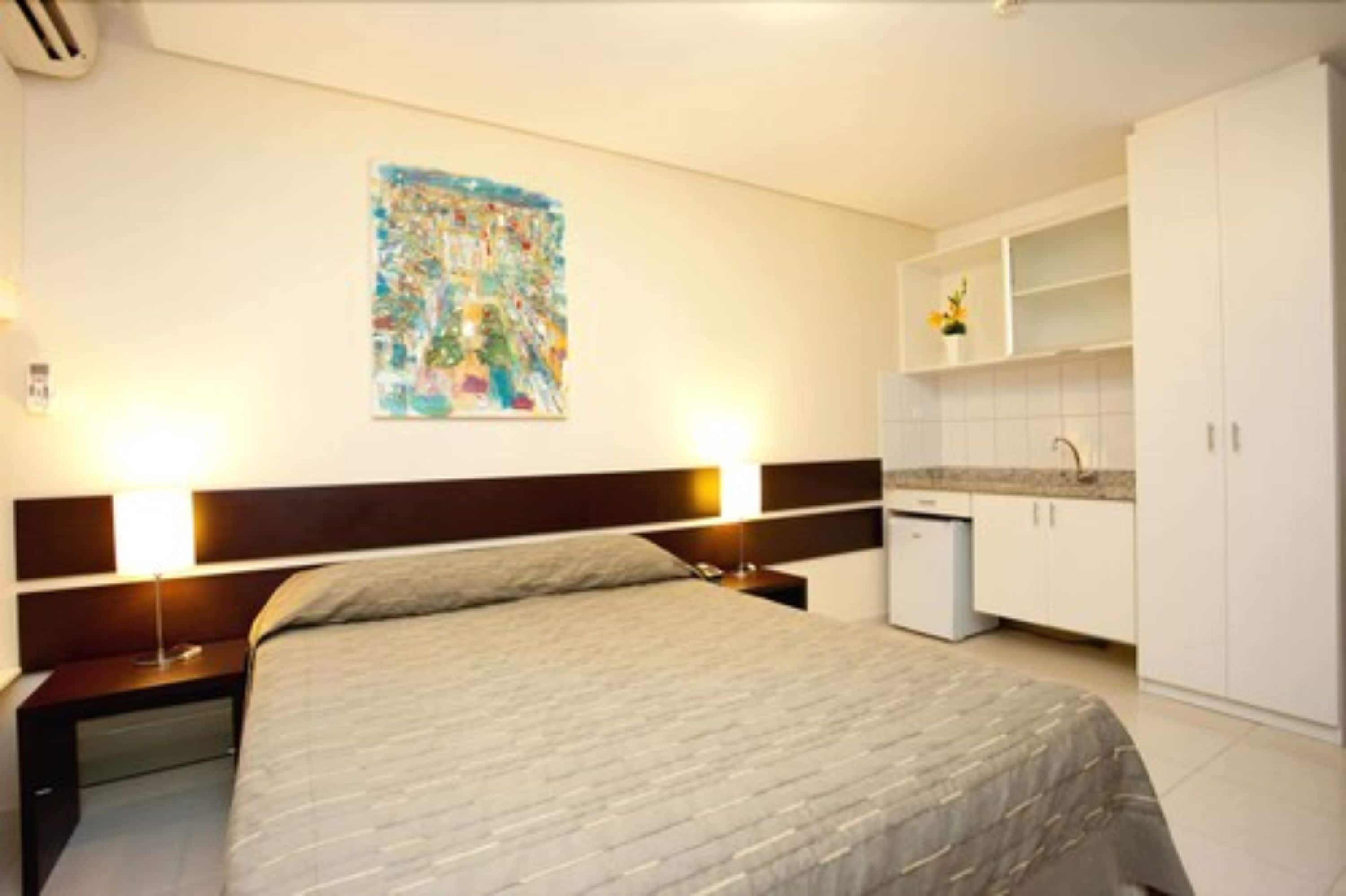 Habitación de hotel con cama, mesitas de noche, un cuadro en la pared y una zona de kitchenette.
