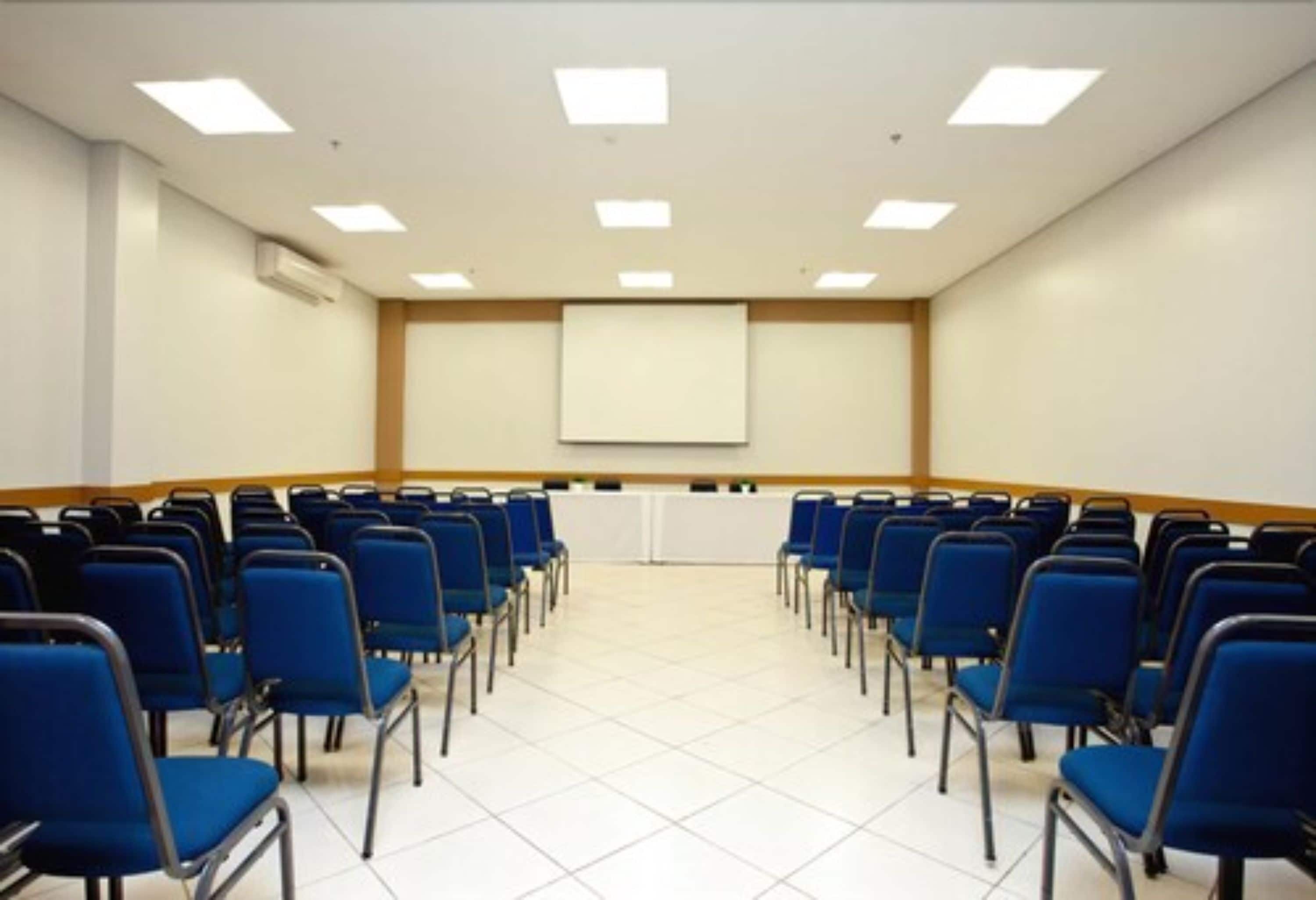 A conference room with rows of blue chairs, a white projection screen, and ceiling lights.