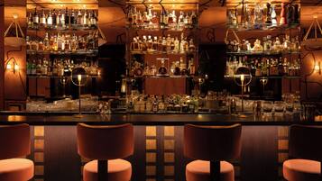 Un espace bar avec de nombreuses étagères remplies de bouteilles, de verres et d’accessoires de bar. On peut voir au premier plan trois tabourets de bar rembourrés.