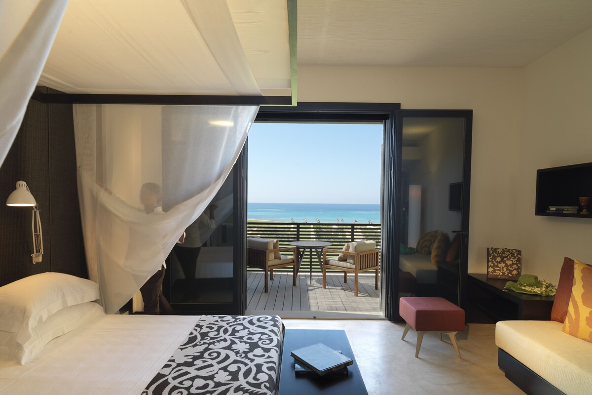 A bedroom with a bed, a bedside table, a laptop, a bedside lamp, a window with a view of the ocean, a balcony with a table and chairs, and a sofa.