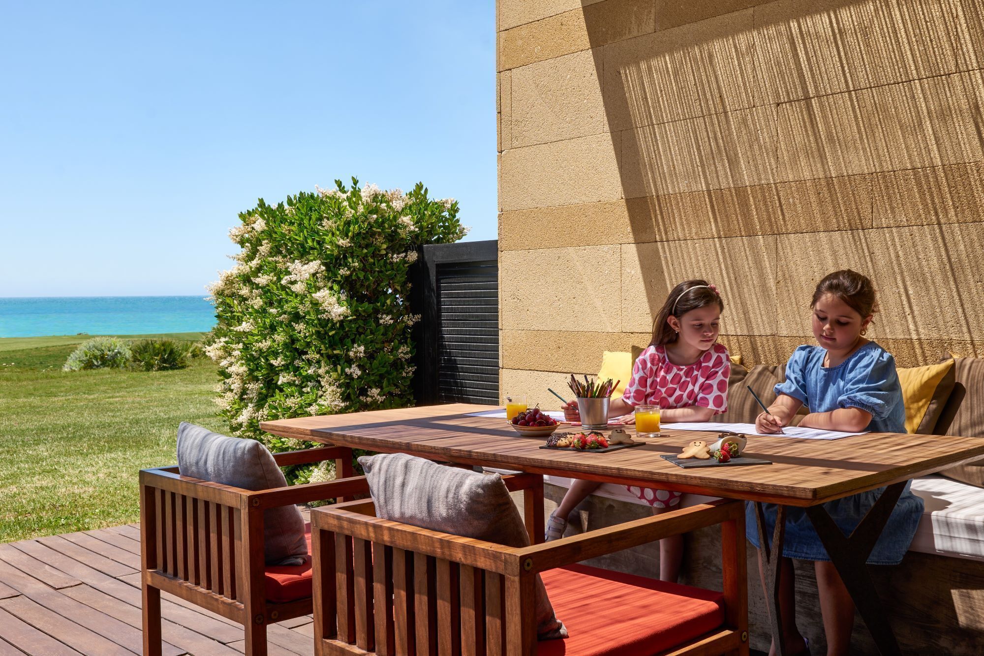 Un espacio para comer al aire libre con vista al océano, mobiliario de madera y una pared de piedra.