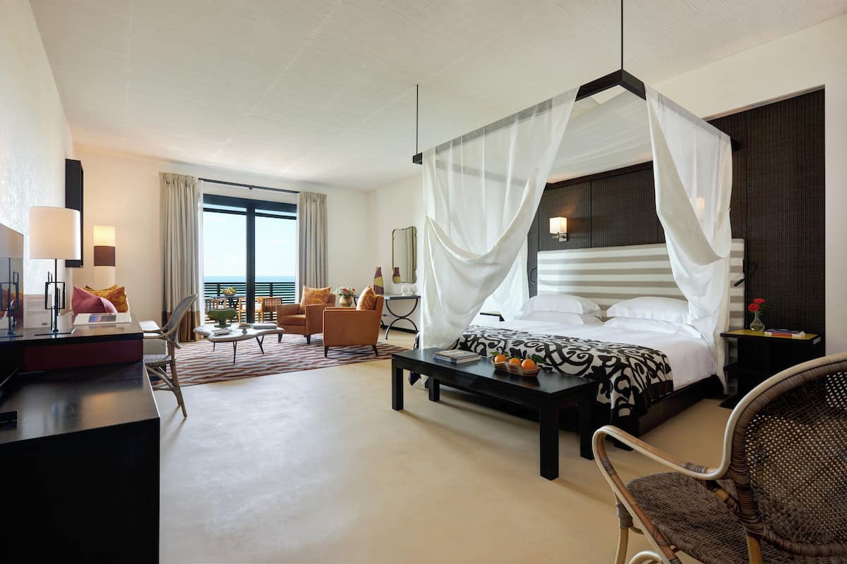 Ein Hotelzimmer mit Himmelbett, Schreibtisch mit Lampe, Stuhl und Meerblick durch große Fenster.