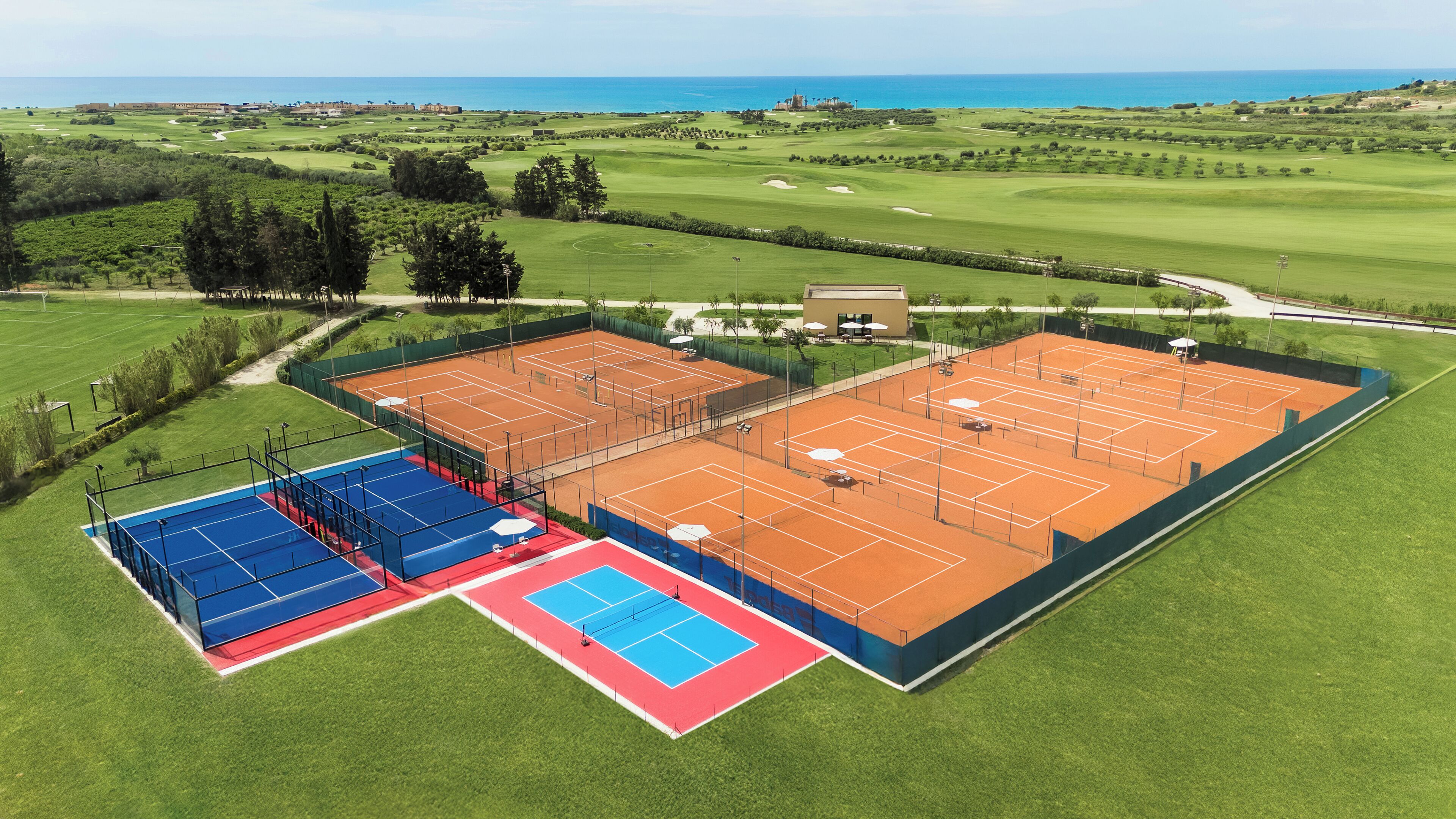 A tennis court complex with multiple courts, a golf course, and a view of the ocean.
