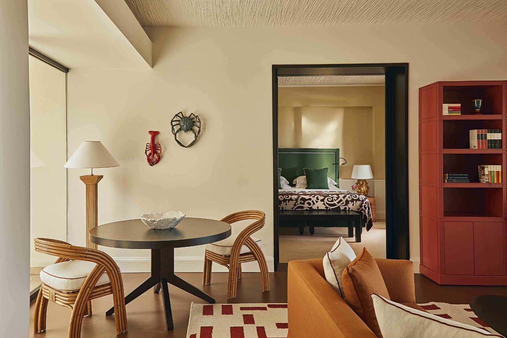 Una sala moderna con una mesa de comedor redonda, sillas de mimbre, un sofá, una estantería roja y una cama con cabecera verde al fondo.