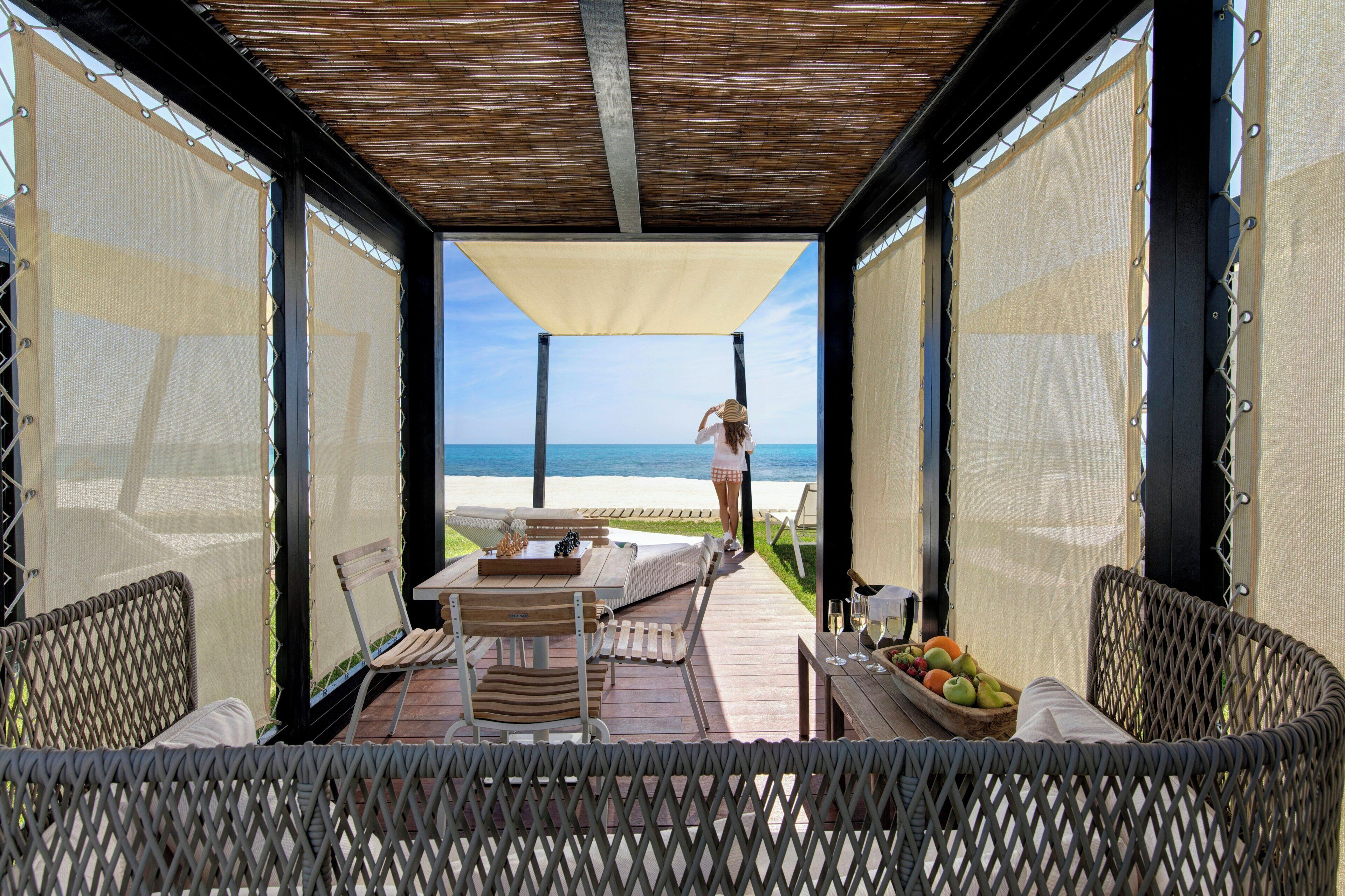 An outdoor seating area with a view of the beach, a table with chess pieces, and a fruit bowl.