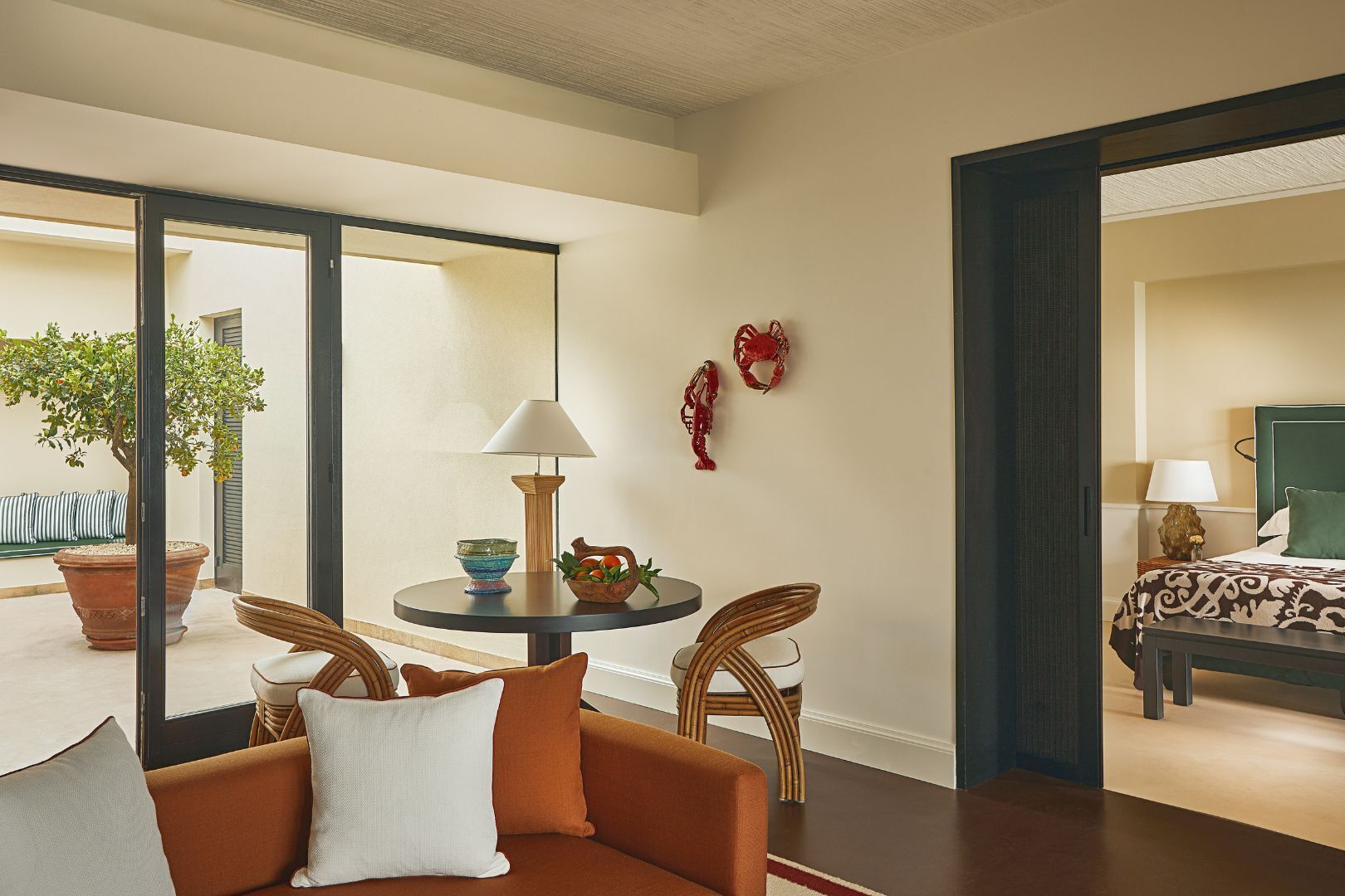 Una habitación de hotel moderna con un sofá, una mesa redonda con lámpara, una planta en maceta y vistas a otra habitación.