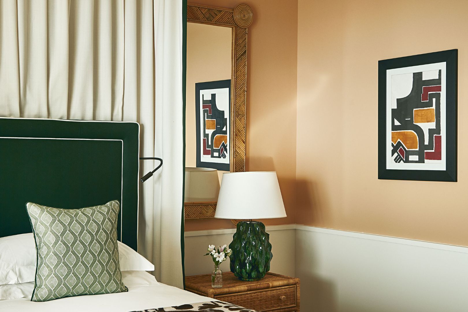 A hotel room with a bed, a nightstand with a lamp, a mirror, and two framed abstract artworks on the wall.