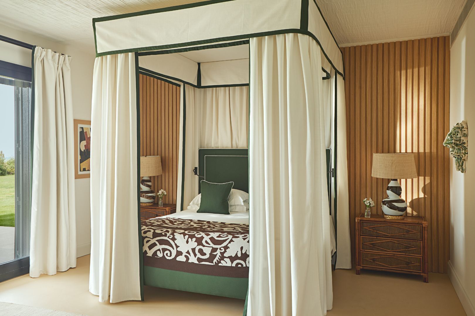 Ein Himmelsbett mit einem gemusterten Bettüberwurf, zwei Nachttische mit Lampen und Blick auf eine grüne Rasenfläche durch eine Glastür.