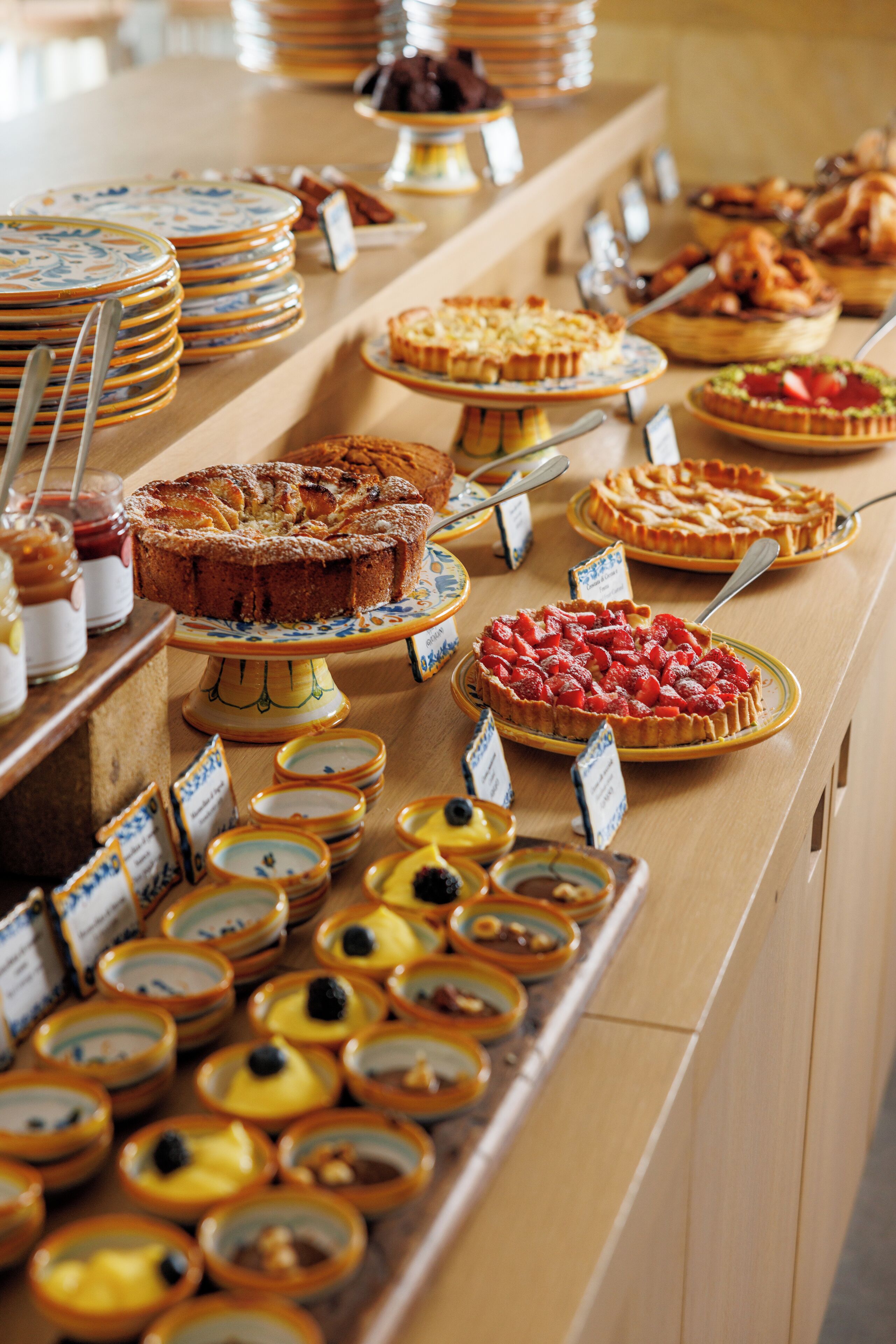 Un buffet con diversi dolci e piatti, tra cui torte, crostate e tartellette di frutta, disposti su un bancone di legno con utensili da portata ed etichette dei prezzi.