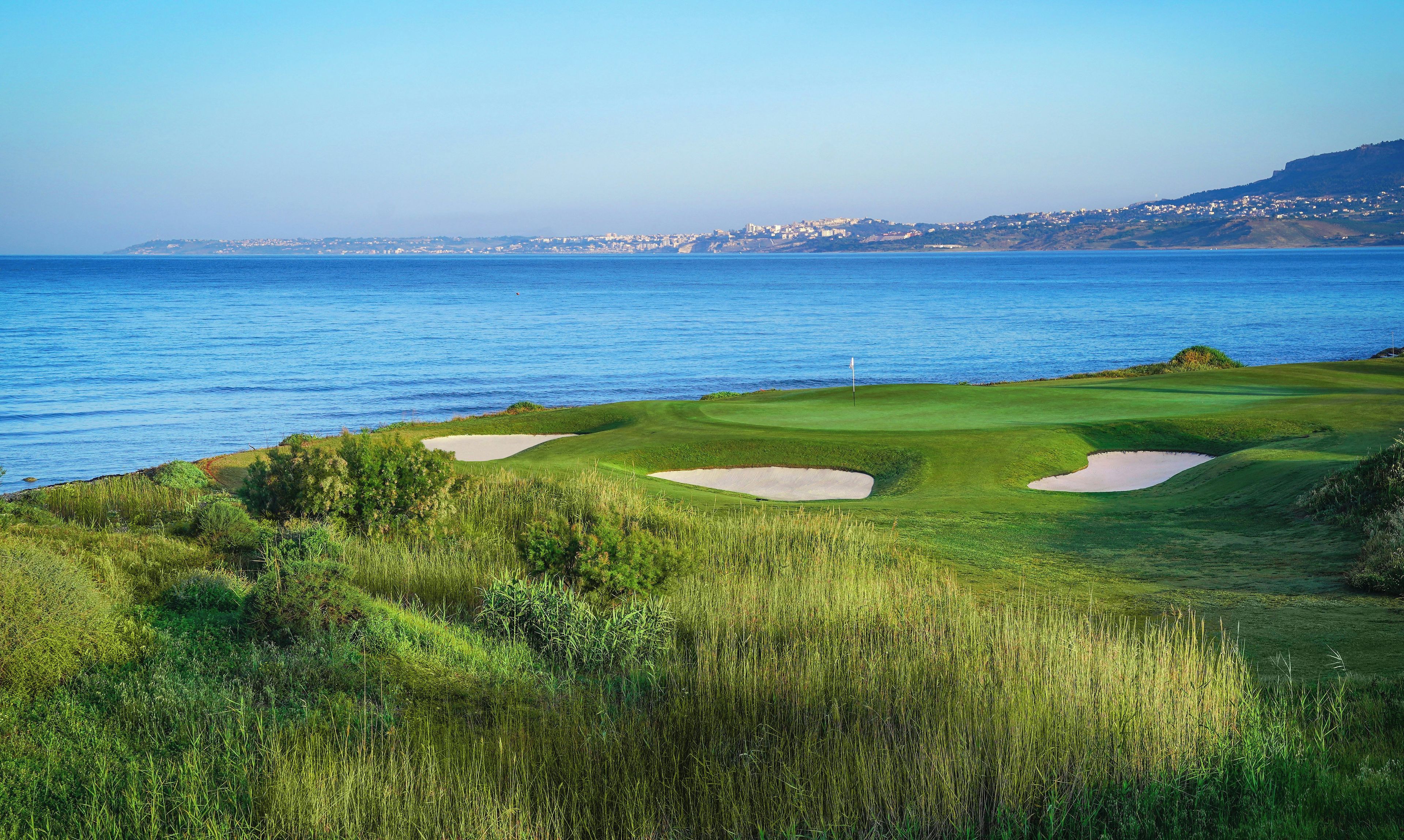 Un parcours de golf situé sur la côte, avec un mât de drapeau, des bunkers de sable et une vue sur la mer et les bâtiments éloignés.