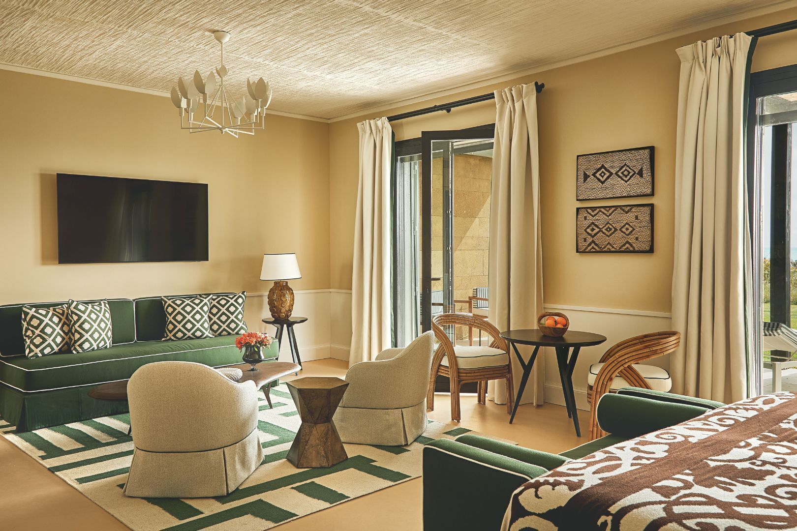 A hotel room with a green sofa, a dining table, a chandelier, and a large TV.
