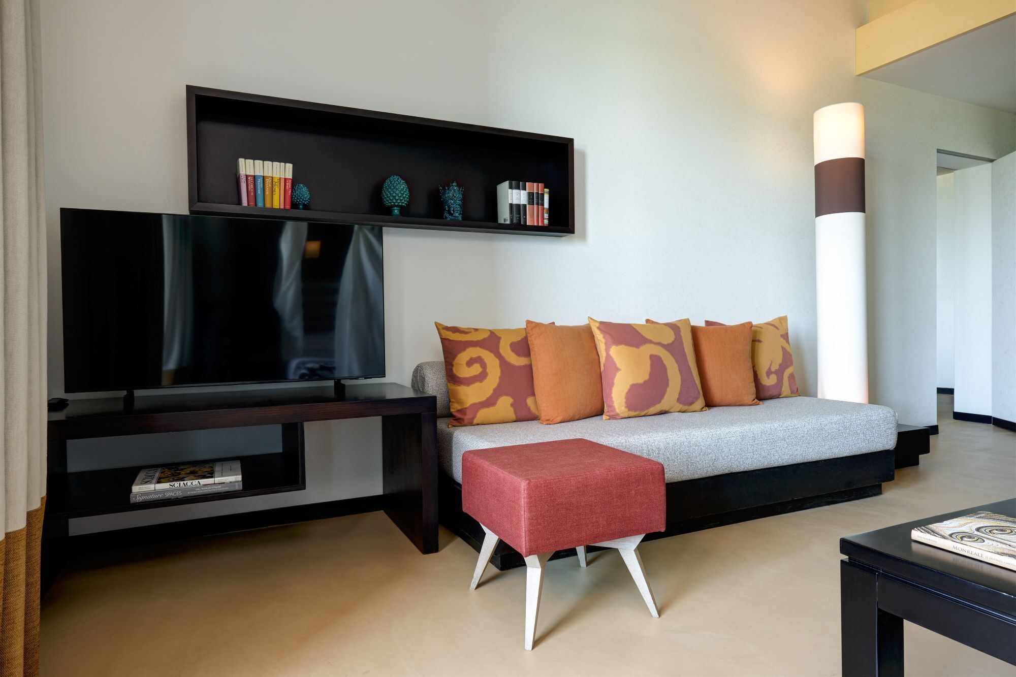 Uma sala de estar moderna com uma TV de tela plana, um sofá com almofadas estampadas, um banquinho vermelho e uma estante com livros e objetos decorativos.