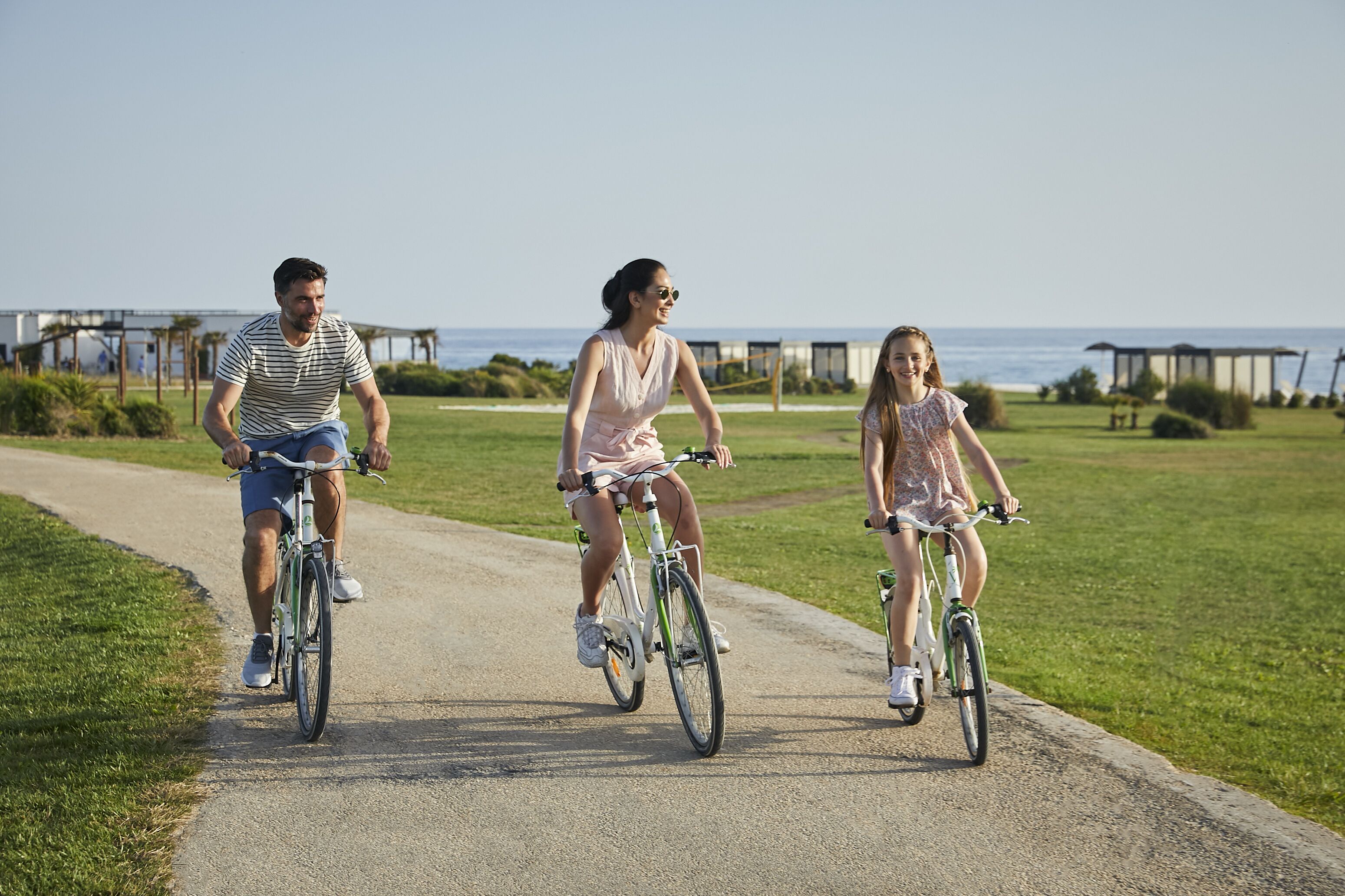 Three people riding bicycles on a paved path with a grassy area and a body of water in the background.
