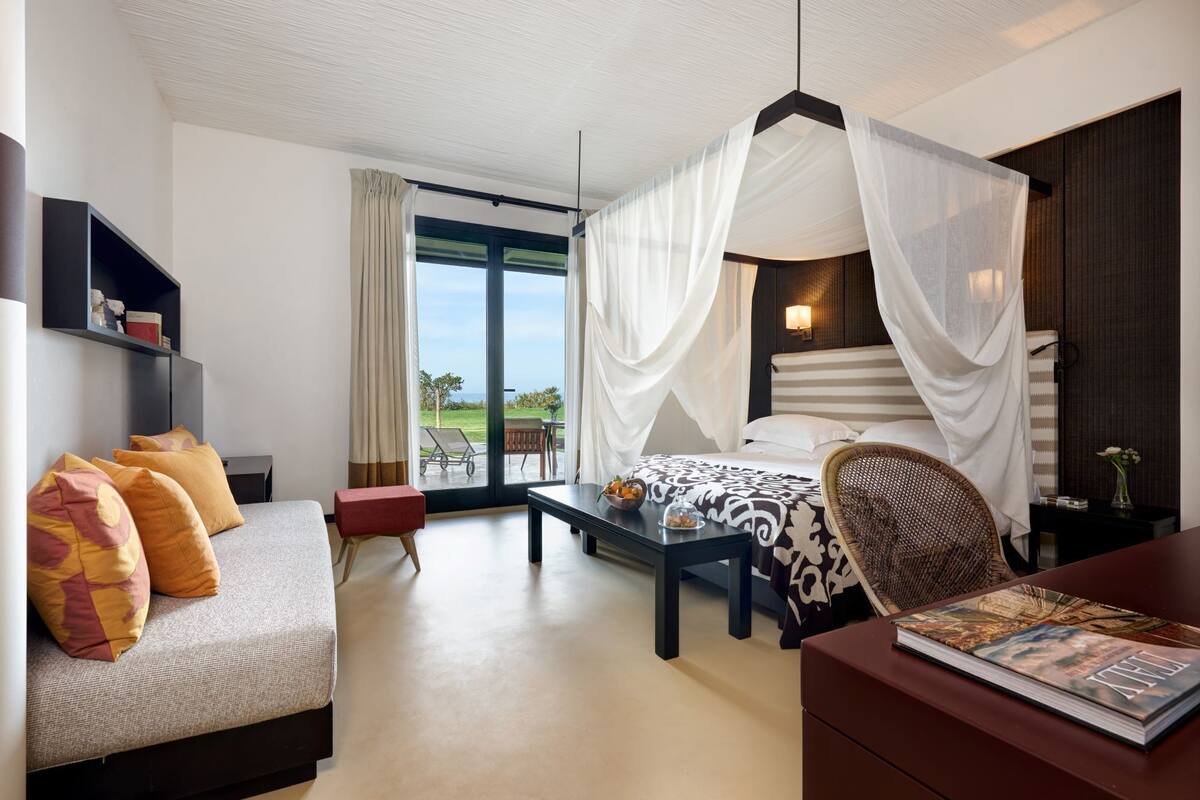 A hotel room with a bed, a sofa, a dining table, and a chair. There is a large window with a view of the outdoors.