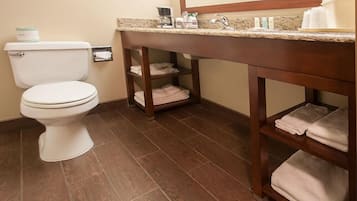 Un baño con inodoro blanco, tocador de madera con lavamanos y espejo encima. Hay toallas en estantes y un cuadro en la pared.
