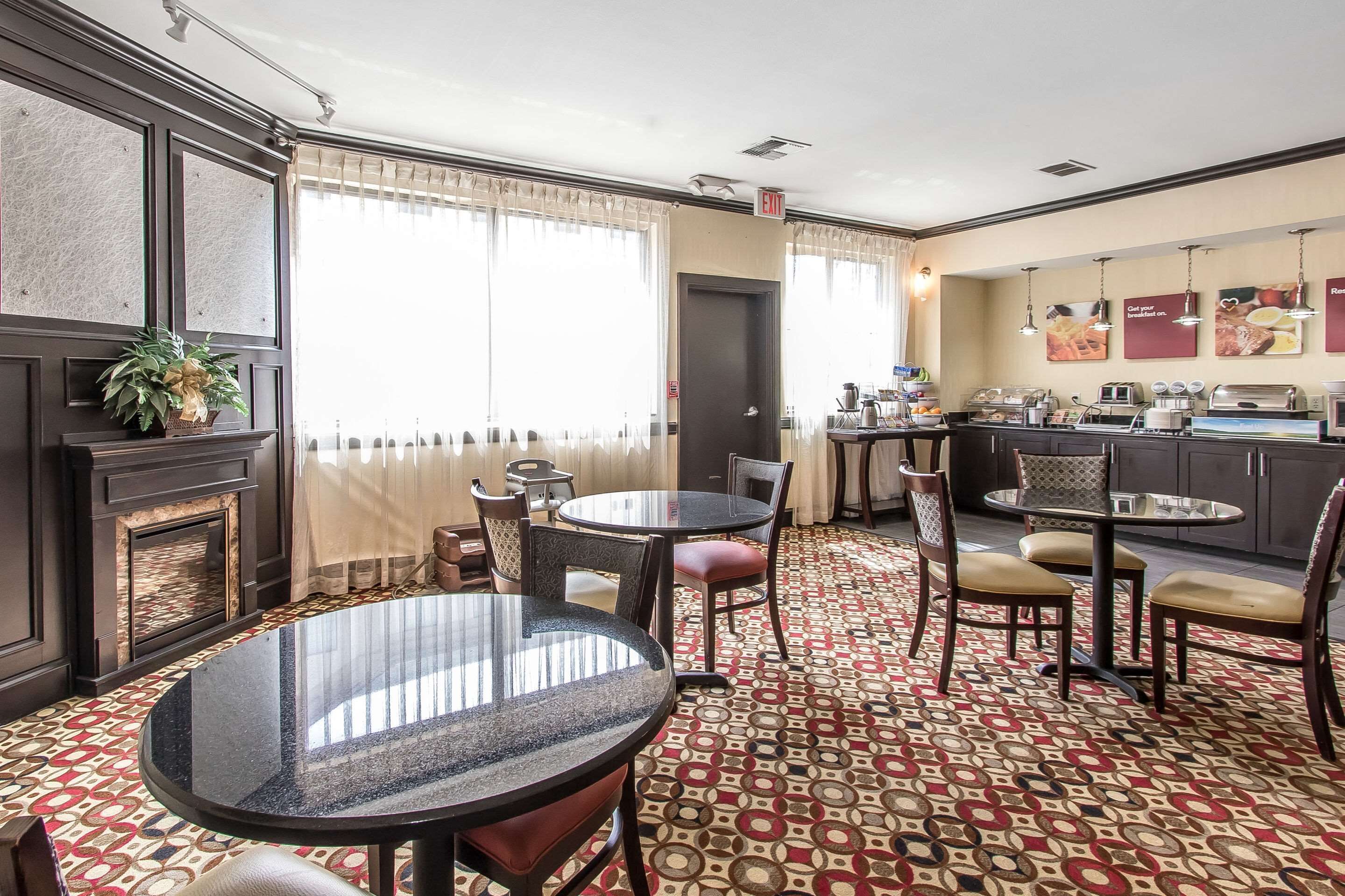 A hotel room with a dining area featuring round tables and chairs, a kitchen area with appliances, and a patterned carpet.