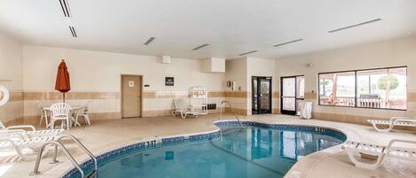 An indoor swimming pool with clear blue water, surrounded by white lounge chairs and a tiled floor.