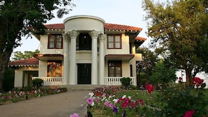 Una casa de dos pisos con una entrada central flanqueada por columnas, techo de tejas rojas y un porche con barandilla blanca.
