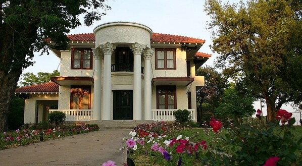 Una casa de dos pisos con una entrada central flanqueada por columnas, techo de tejas rojas y un porche con barandilla blanca.