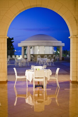 An outdoor dining area with tables and chairs, an archway, and a building in the background. - Kipriotis Panorama Hotel & Suites by Anayia All Inclusive Resorts (Kos)
