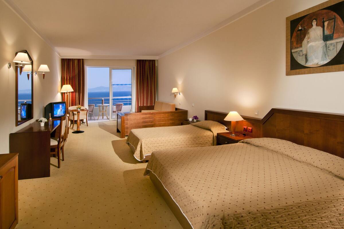 Une chambre d’hôtel avec deux lits, un bureau avec une télévision, un coin repas et une vue sur la mer.