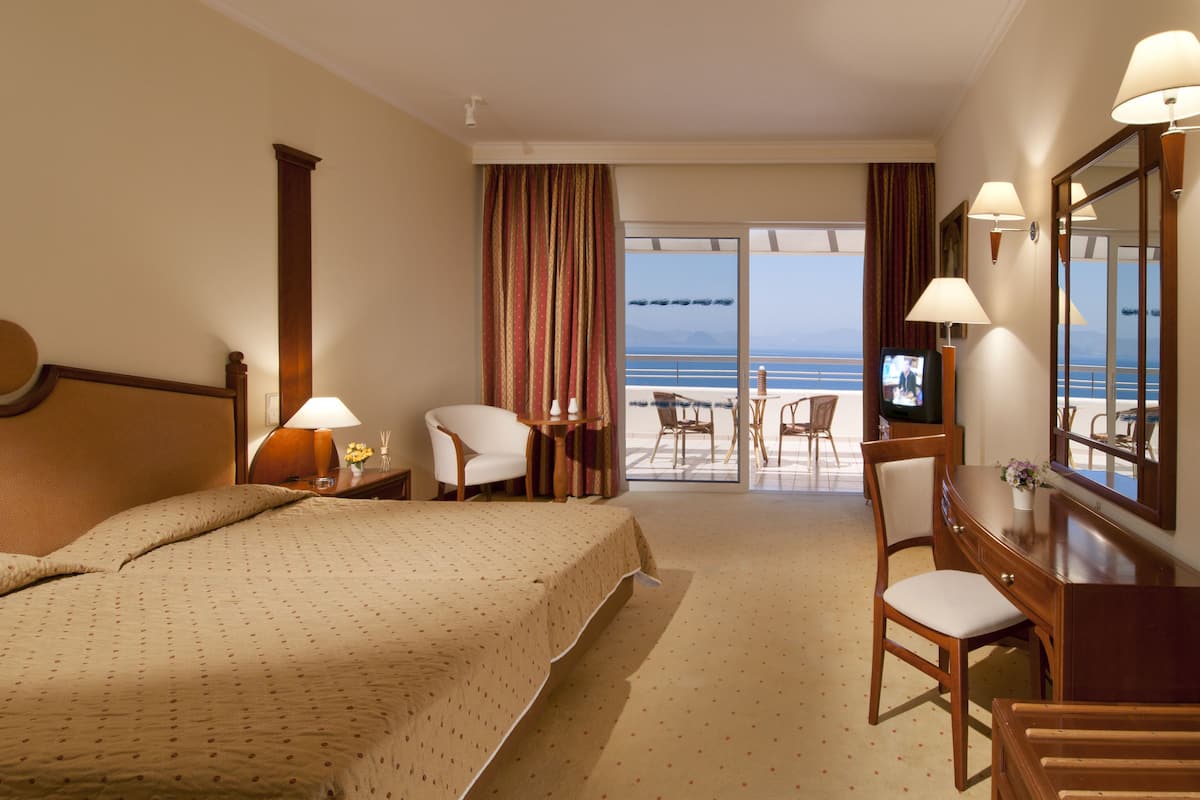 Une chambre d’hôtel avec un grand lit, un bureau avec une chaise, une télévision et une vue sur la mer par les fenêtres.