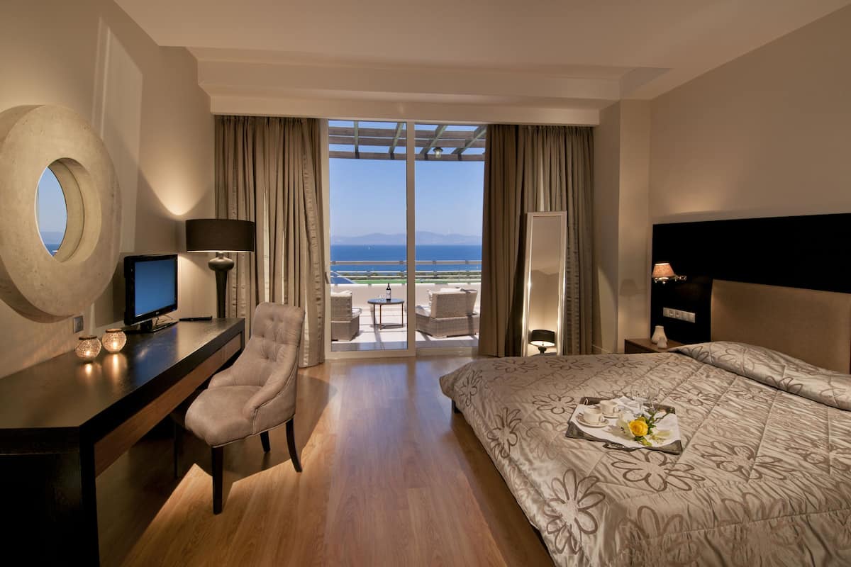 Une chambre d’hôtel équipée d’un lit, d’un bureau, d’une chaise, d’une télévision et offrant une vue sur la mer grâce à de grandes fenêtres.
