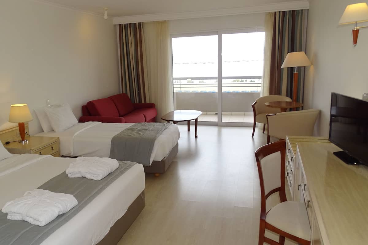Une chambre d’hôtel avec deux lits, un canapé rouge, une petite table, un téléviseur et un balcon avec des rideaux.