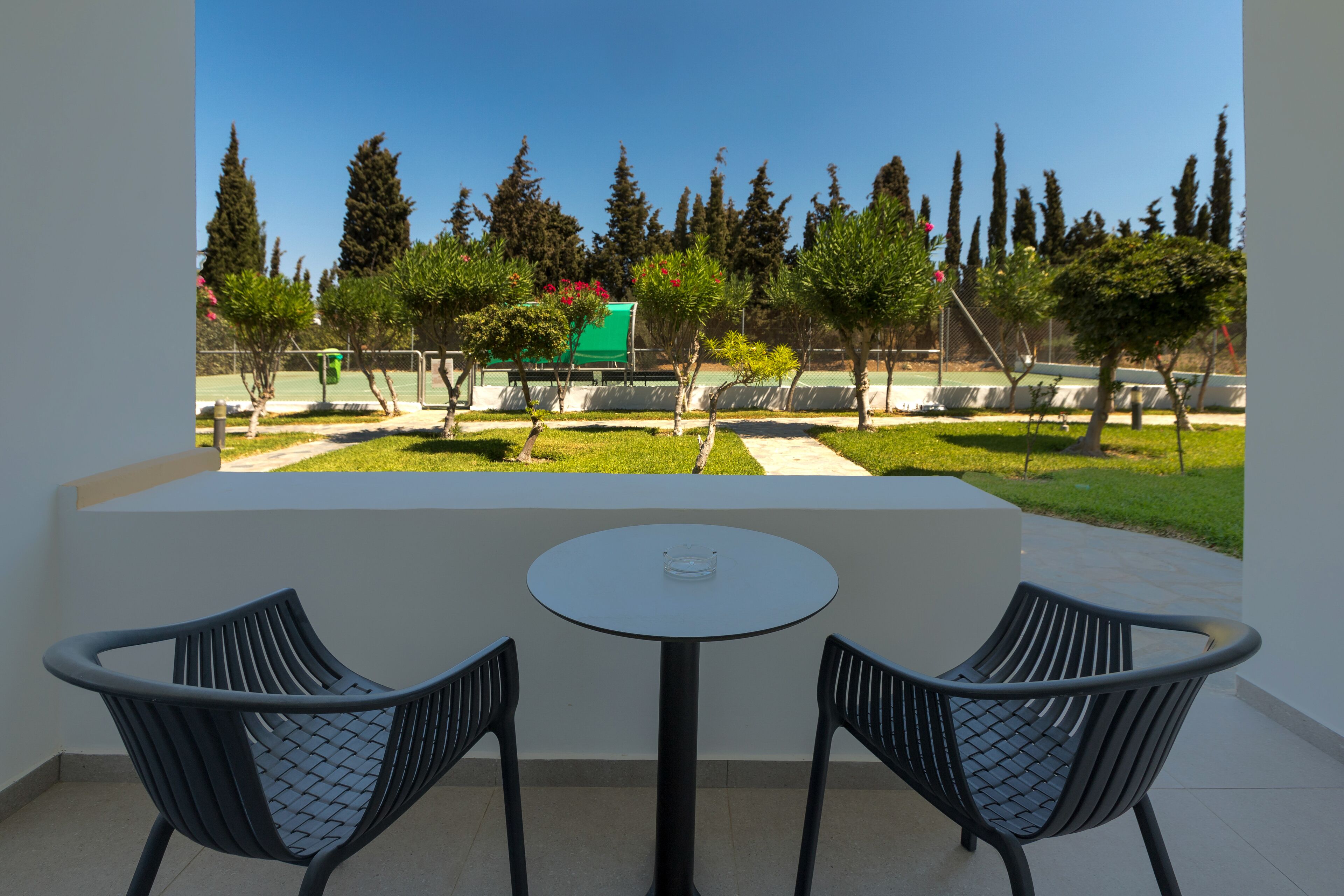 A balcony with two black chairs and a small table, overlooking a well-maintained garden with trees and a tennis court.