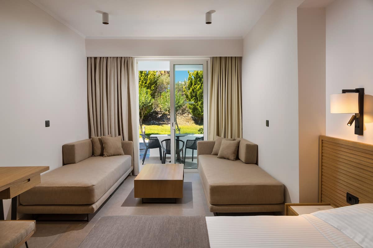 A hotel room with two sofas, a coffee table, a bed, and a view of a garden through a glass door.