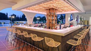 An outdoor bar area with a central wooden structure, bar stools, and a variety of bottles. - Hotel Platanista (Kos)