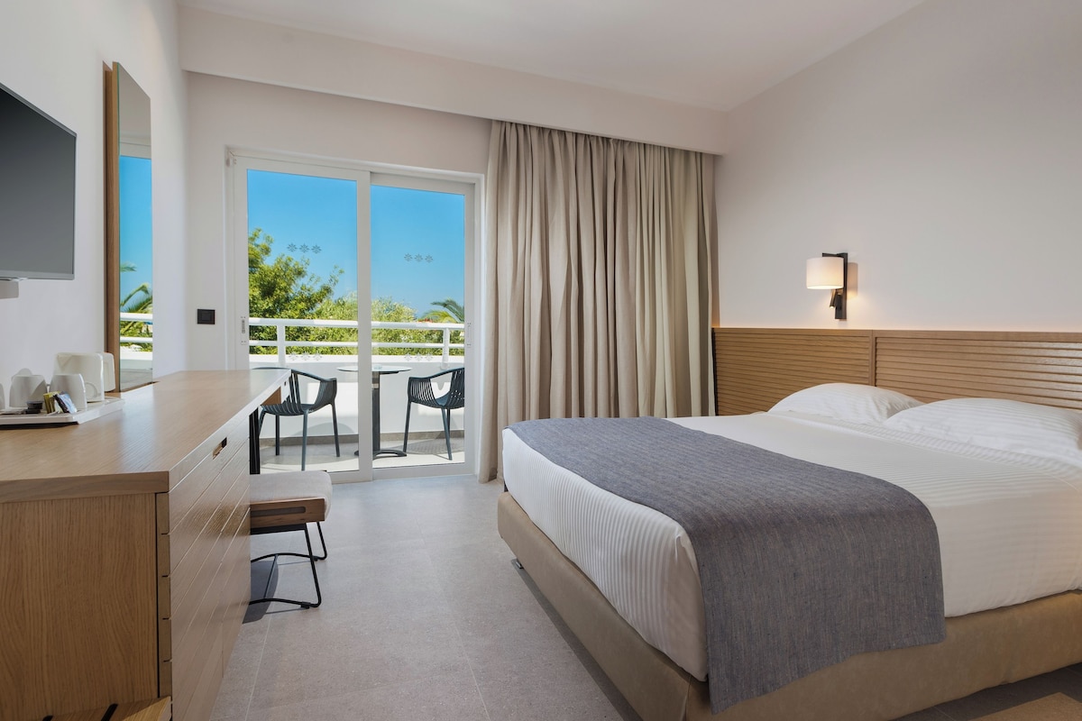 Habitación de hotel con cama, mesita de noche, televisión y un balcón con sillas.