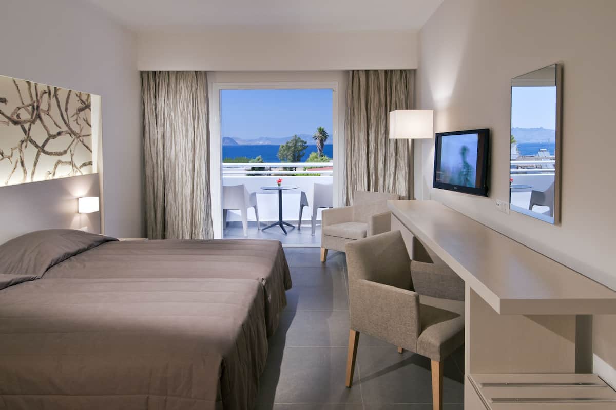 A hotel room with a bed, a desk, a chair, a TV, and a view of the sea.