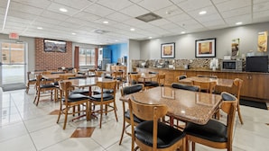 Un grand espace de salle à manger avec plusieurs tables et chaises, un coin cuisine doté d’un comptoir et d’appareils électroménagers, et un mur de briques orné d’un tableau encadré.