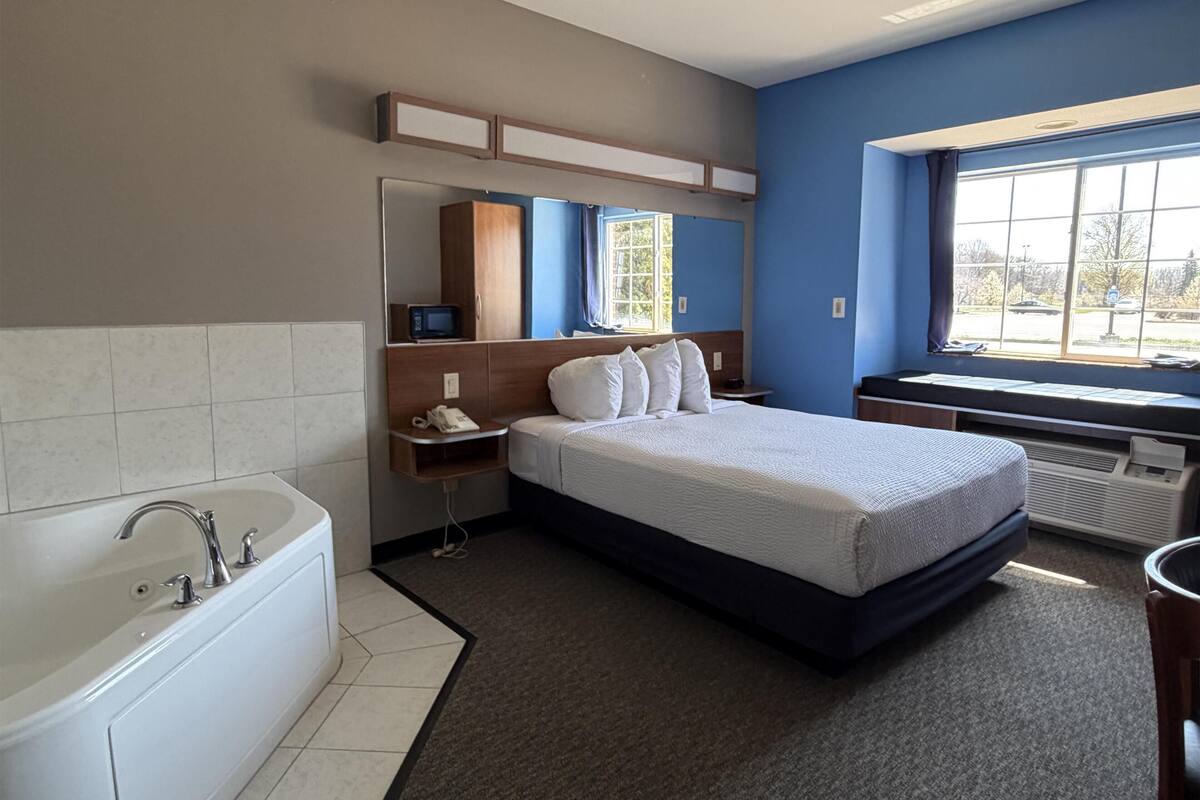 Une chambre d’hôtel avec un lit, une salle de bain avec une baignoire, un bureau avec un ordinateur, une fenêtre donnant sur un paysage agréable et un miroir au-dessus du lit.