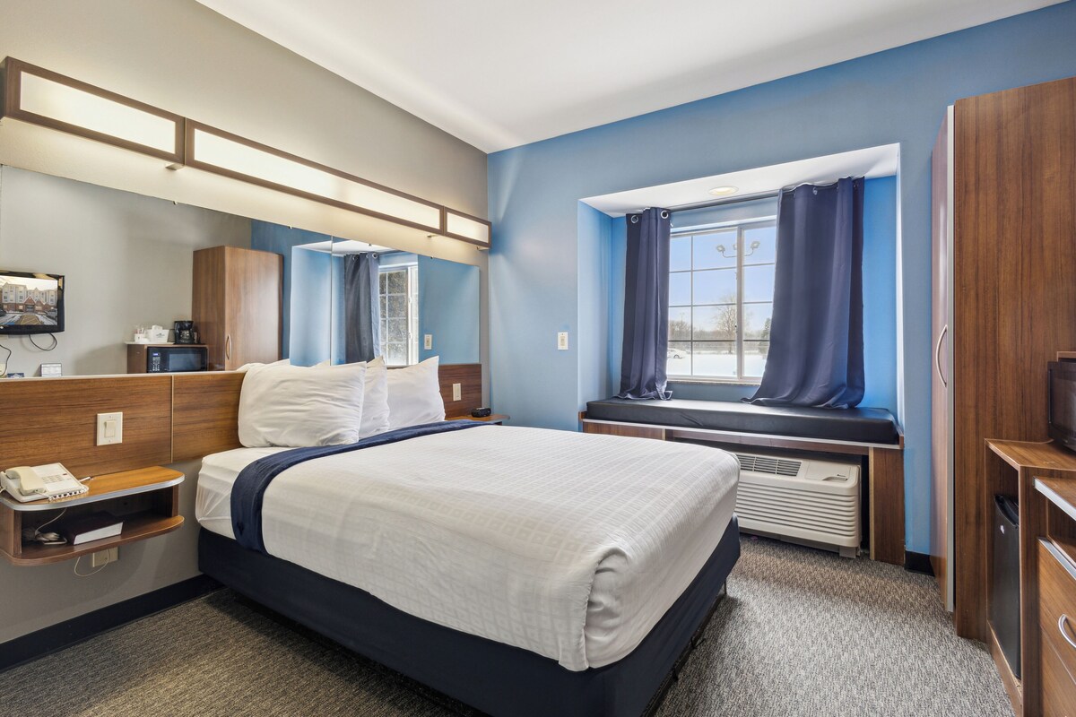 A hotel room with a large bed, a bedside table with a phone, a window with curtains, a built-in bench, and a wall-mounted TV.
