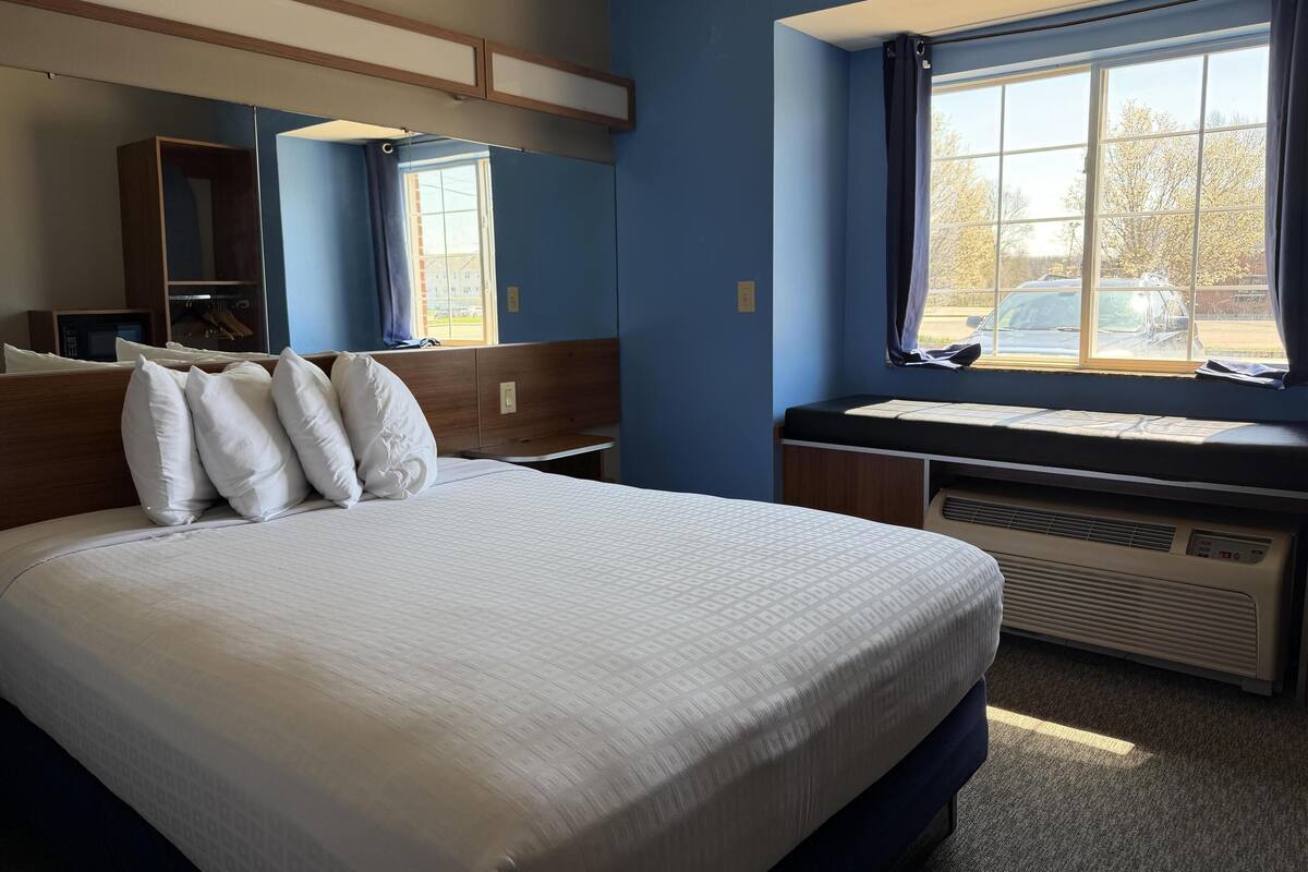 Une chambre d’hôtel avec un grand lit, un bureau, une fenêtre avec des rideaux, un miroir et un climatiseur fixé au mur.
