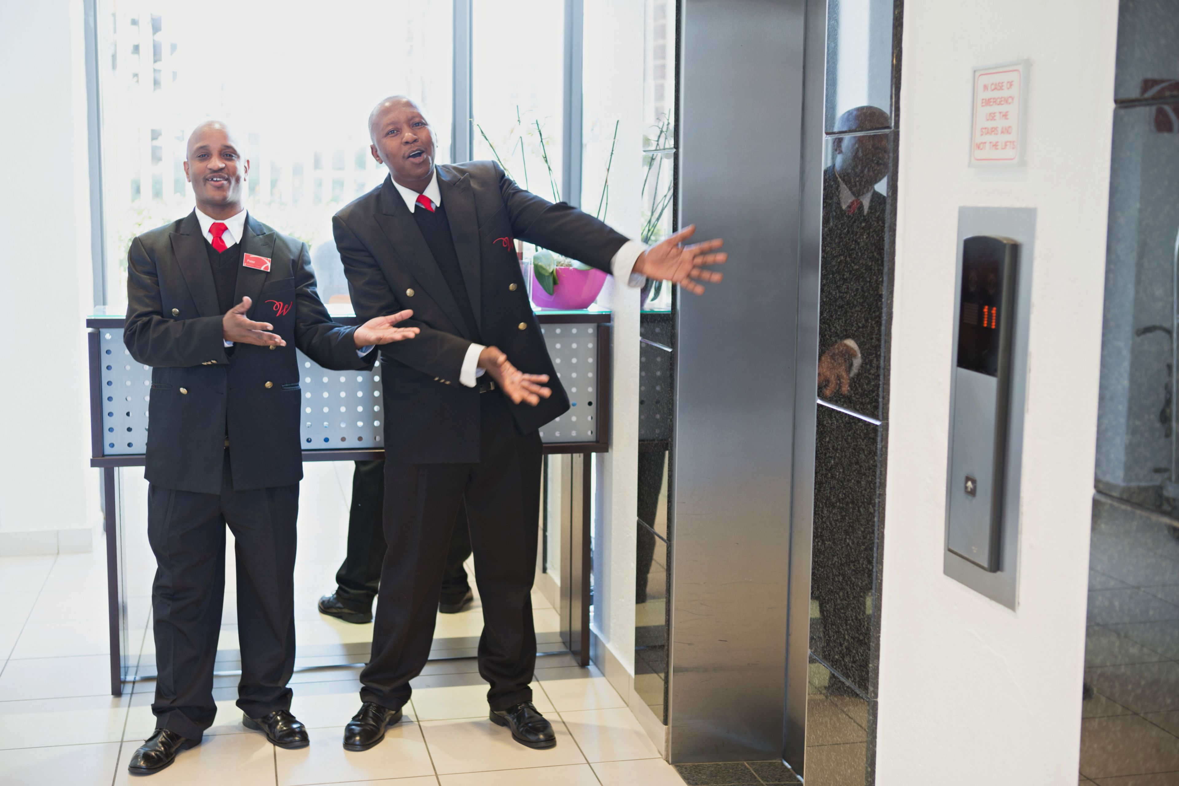 Two men in suits standing in an elevator with a potted plant and a digital display on the wall.