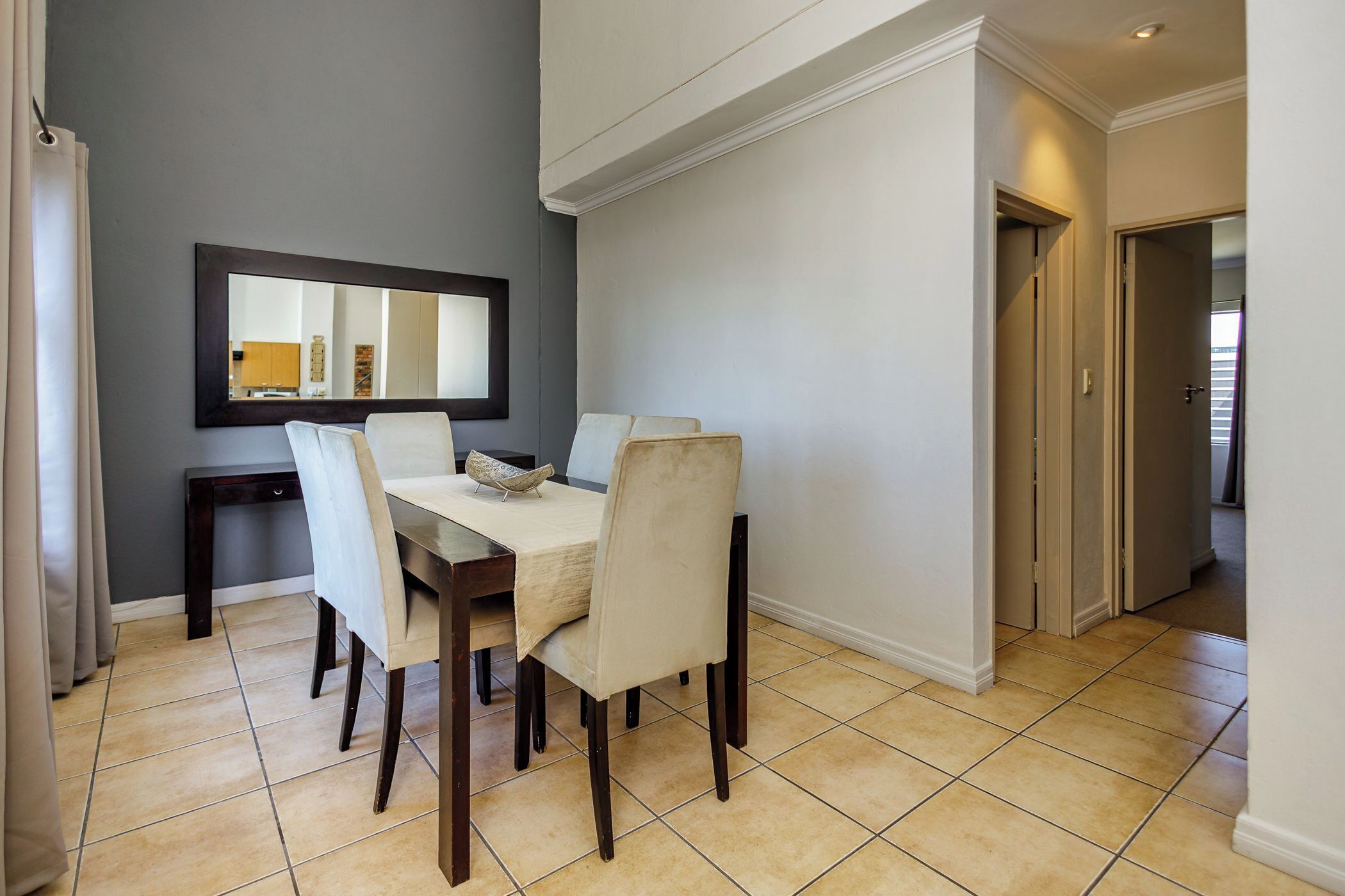 Een eethoek met een tafel en stoelen, een spiegel aan de wand en een deur naar een andere kamer.