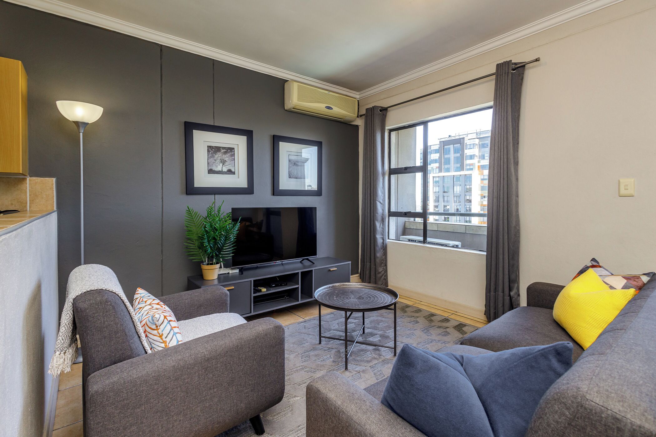 A modern living room with a flat-screen TV, grey sofas, a coffee table, and a large window with curtains.