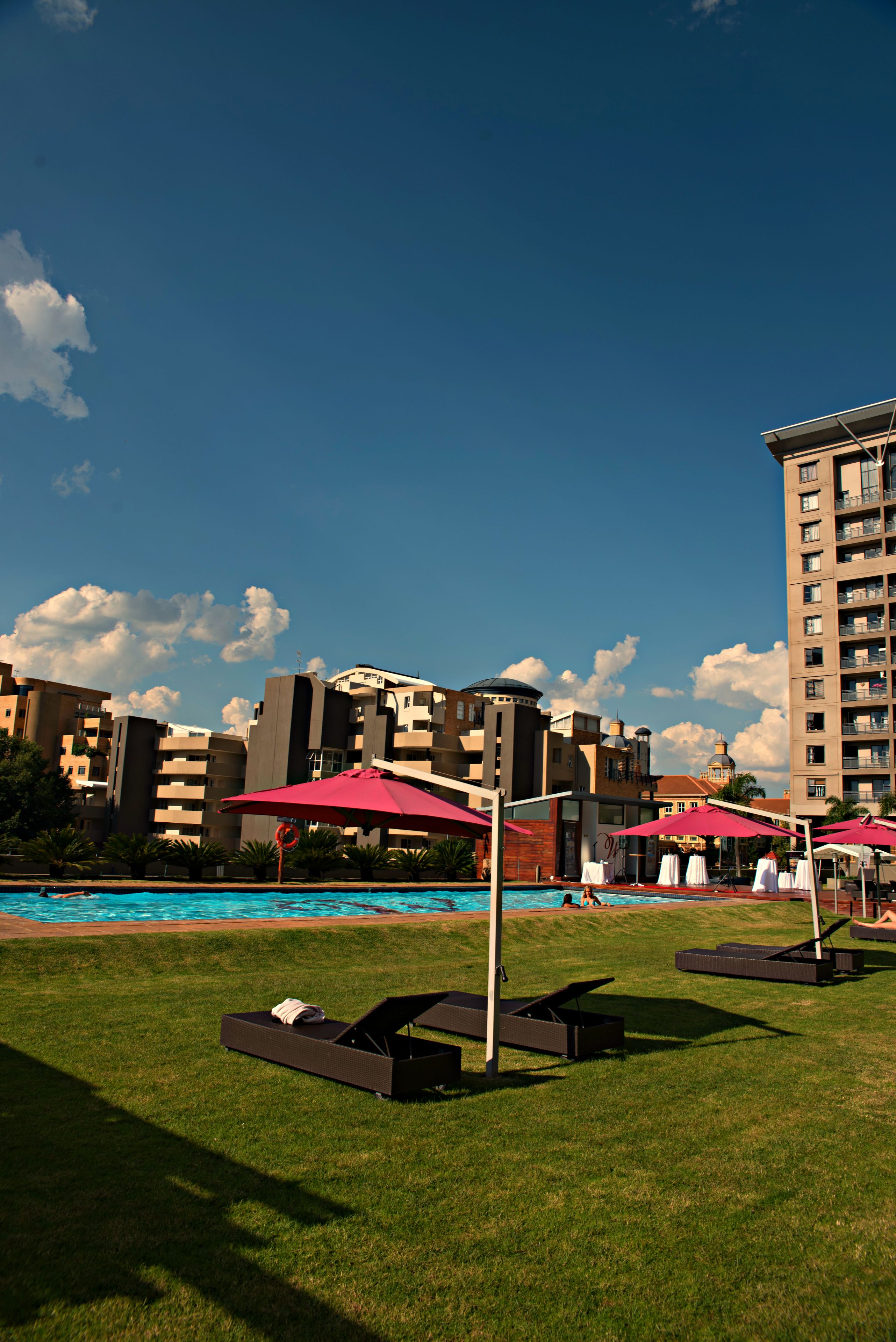 A poolside area with lounge chairs and umbrellas, a clear blue sky, and a building in the background.