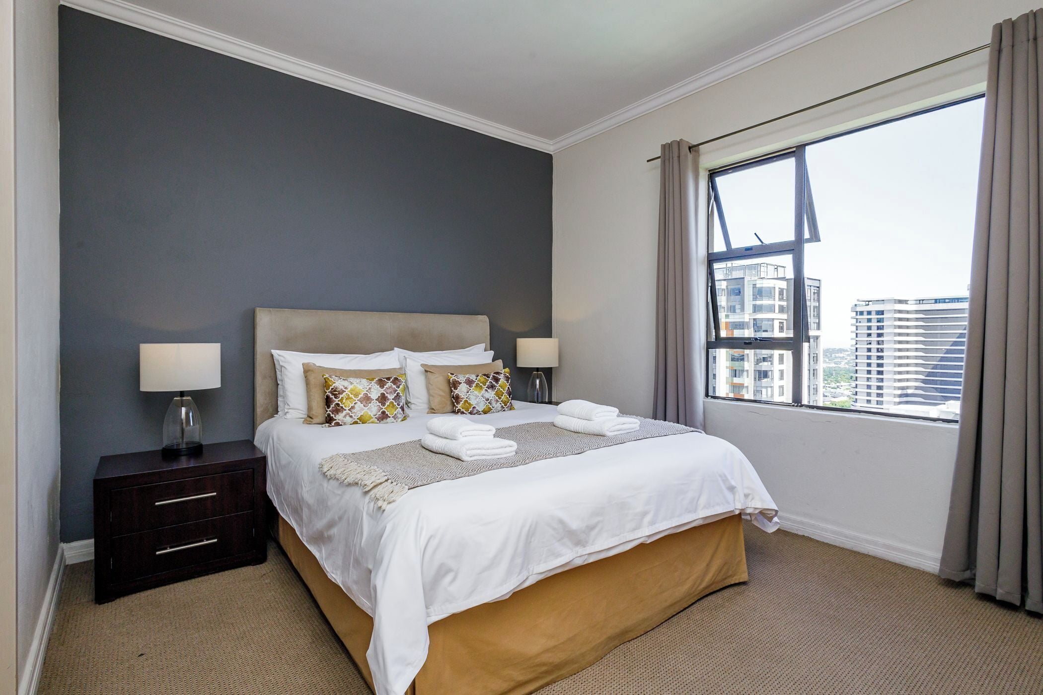 A hotel room with a large bed, two bedside tables, a window with a view of buildings, and a dark-colored wall.