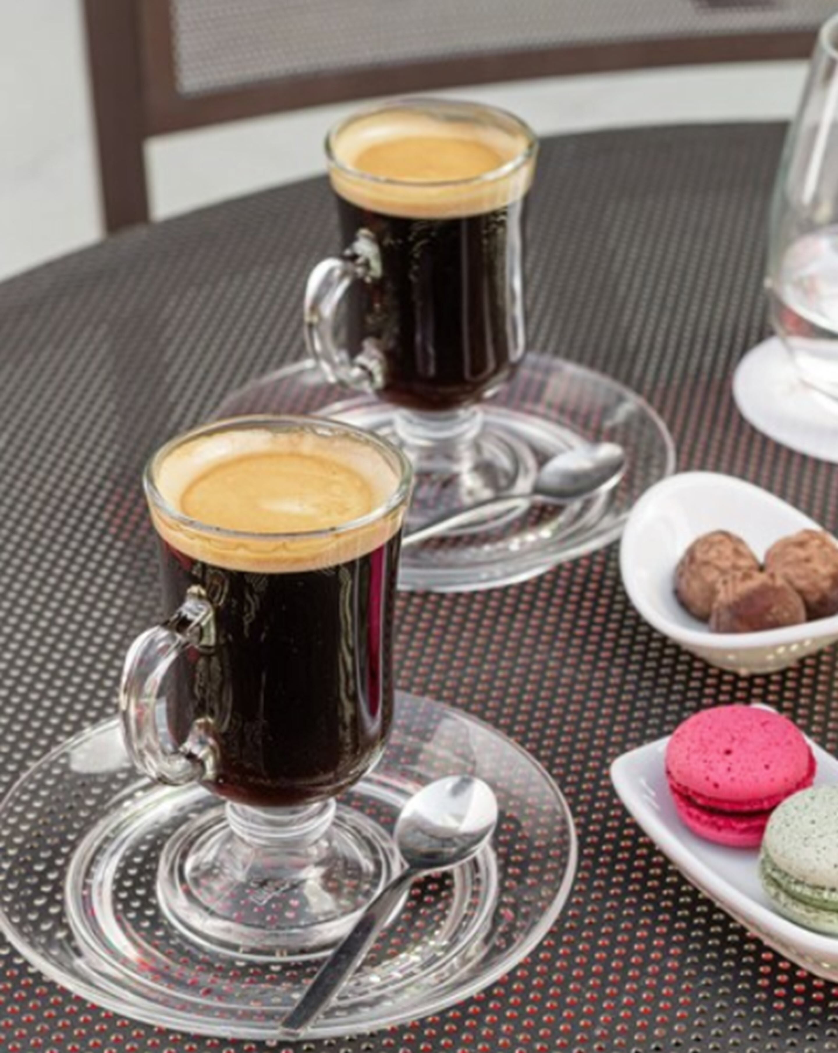 Two dark-colored glasses of a beverage on a table with a metal spoon and a plate of cookies.