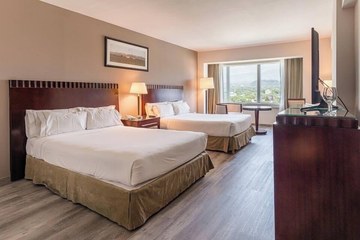 A hotel room with two beds, a desk, a chair, a TV, and a window with a view.