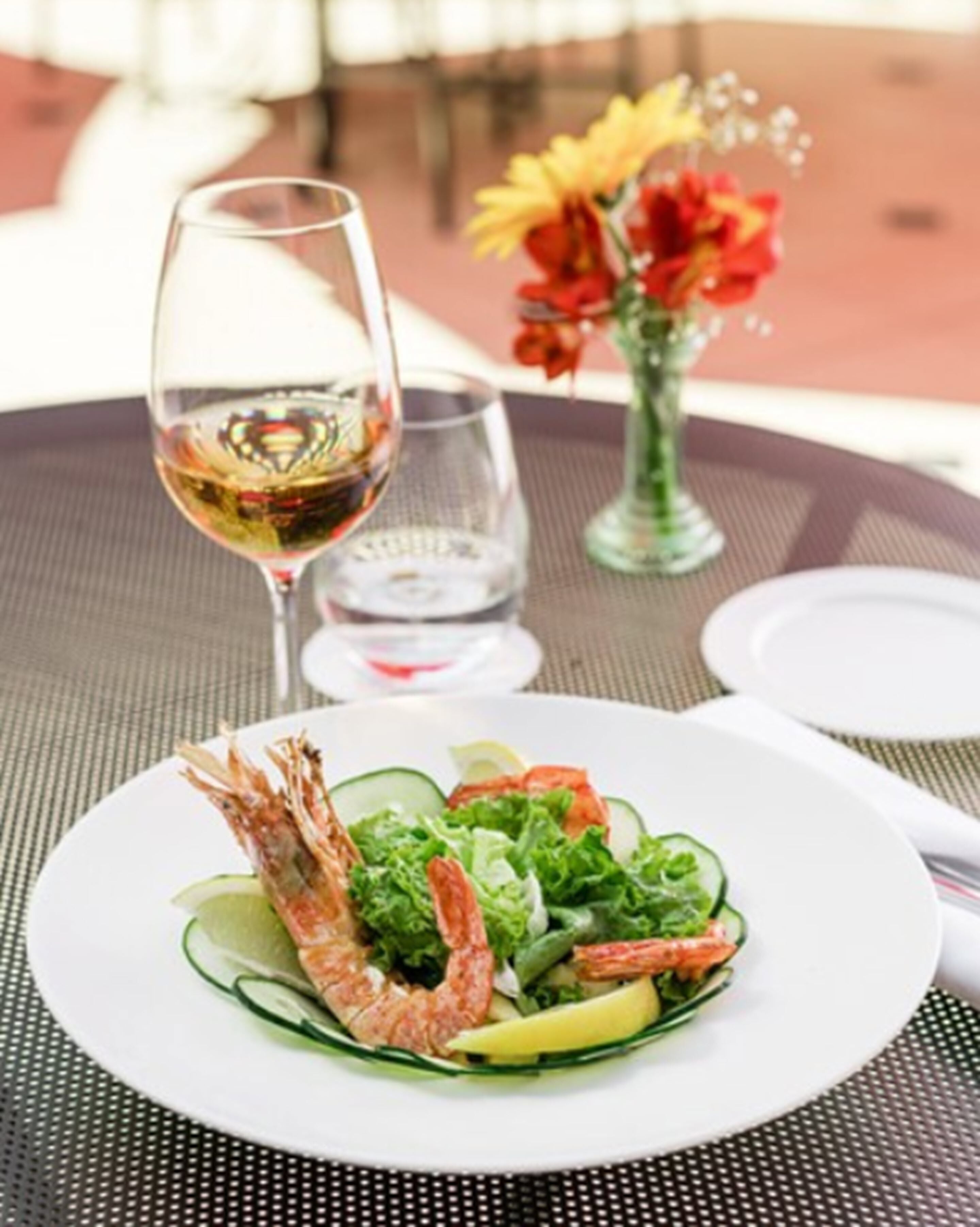 A dining table with a plate of shrimp salad, a glass of white wine, and a vase with flowers.